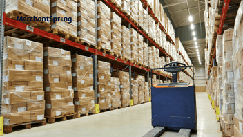 Large-scale warehouse with stacked pallets of boxed inventory and an industrial pallet mover, representing Amazon wholesale as a scalable 1P operating model