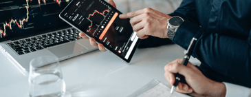 Two business professionals analyzing data charts on a tablet and laptop, reviewing market trends and forecasts in a modern office setting